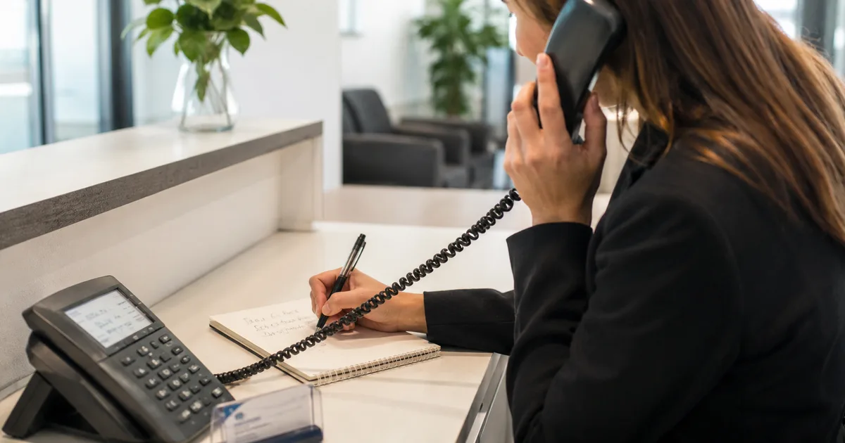 Resepsiyon masasında çalışan bir kişi telefon ahizesini tutarken not defterine arayanın talebini yazıyor